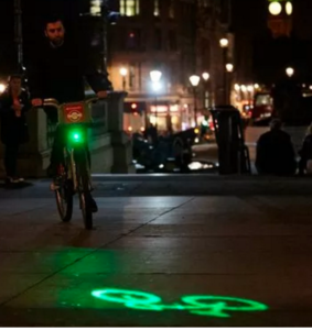 Night scene of cyclists beaming green image of a bicycle meters ahead onto pavement.