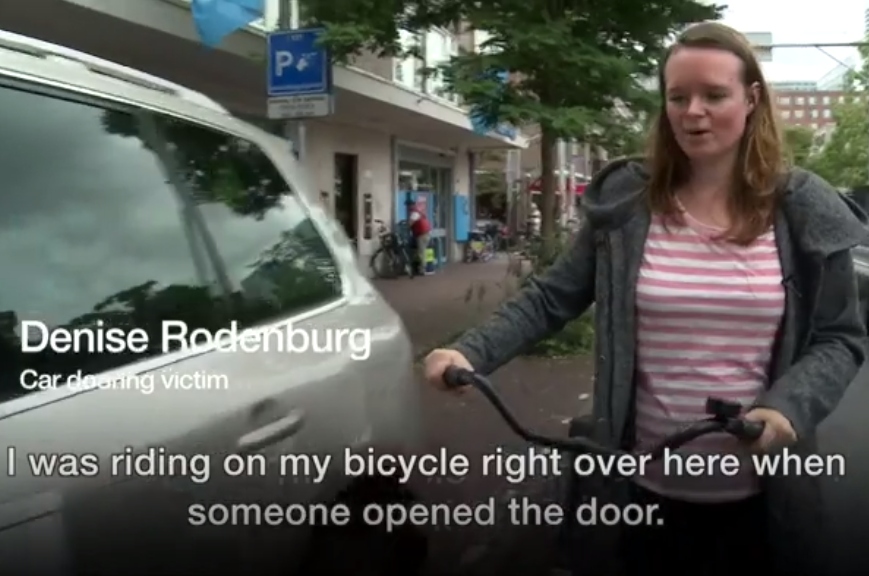 Young woman walks her bicycle beside a parked car, tells how she was doored, injured - follows video re-enactment of her crash / accident hitting carelessly opened car door. This sets up the short educational video How to do the Dutch Reach by the Royal Society for the Prevention of Accidents UK.