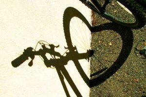 Photo: Bike shadow on wall and adjacent ground, shows hand bars, stem, front wheel & fork. Shadow of wheel bends at ground, forming heart shaped front wheel shadow.  Unknown photographer, found on Google search of bike + heart