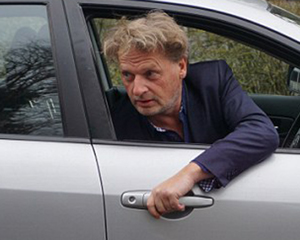 Screen shot of dodgy tabloid reporter's mock "dutch reach', reaching out window to open car door, to discredit NL method either for sensation or in defense of motorist bias against cyclists.