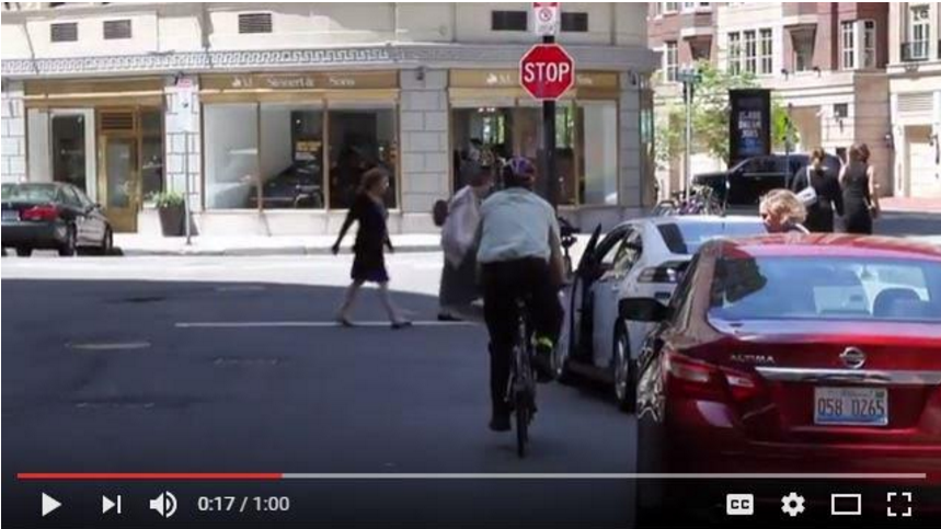 Screen shot from Massachusetts RMV / DOT driver & bicycle safety video on Dutch Reach method. Photo shows cyclist riding along side parked cars. Caption reads: "Because it is difficult for a cyclist to see if there is a driver in each individual car, doors that open in front of their paths can be totally unexpected and forcing the bicyclist to stop on a dime."