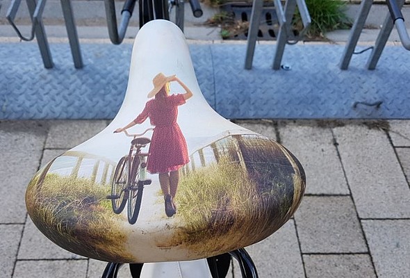IWomans bicycle saddle with painted image of woman walking along side a bike, holding hat on head.